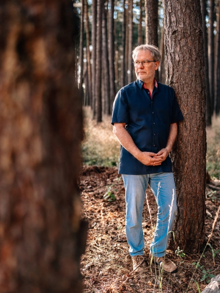 Frank Schmitz steht mit ineinandergelegten Händen im Wald an einem Baum