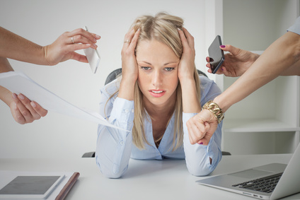 Eine Überforderte Business-Frau sitzt an einem Schreibtisch und Hände halten Ihr Handys und Uhren hin